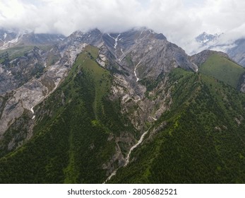 High angle view looking toward steep jagged rocky peaks covered with patches of snow and dense coniferous forest slopes