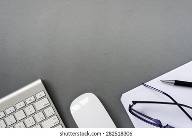 High Angle View Of Mac Computer Keyboard And Mouse With Paper, Pen And Eyeglasses On Grey Desk With Ample Copy Space