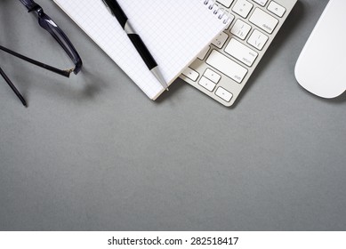 High Angle View Of Mac Computer Keyboard And Mouse With Paper, Pen And Eyeglasses On Grey Desk With Ample Copy Space