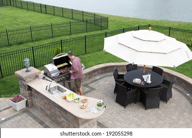 High angle view of a man cooking meat on a gas BBQ standing in the sunshine on a paved outdoor patio at the summer kitchen preparing for guests with a table and chairs with a garden umbrella alongside