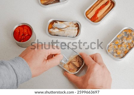 high angle view of a man opening a can of frigate tuna fillets at a counter, where there are some open cans with different food, such as sardines, anchovy fillets, cockles or roasted red bell peppers