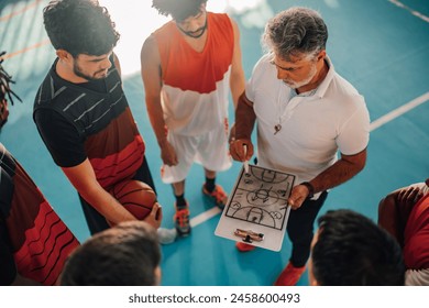 High angle view of a mature confident male coach discussing game tactics with his multiracial basketball team using a coach clipboard and explaining game plan and drawing defense. Copy space. - Powered by Shutterstock