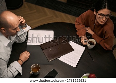 High angle view of middle aged Caucasian couple reviewing divorce papers and wedding rings, sitting apart in conflict. Useful for legal services, separation counseling, family law
