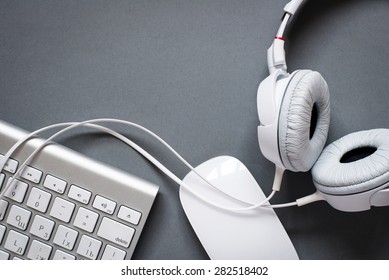 High Angle View Of Modern White Audio Headphones With Cord, Mac Computer Keyboard And Mouse On Grey Desk Background With Copy Space