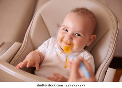 High angle view of mother feeding cheerful baby boy with porridge, baby sitting in high chair all messy and staied smiling and eating - Powered by Shutterstock