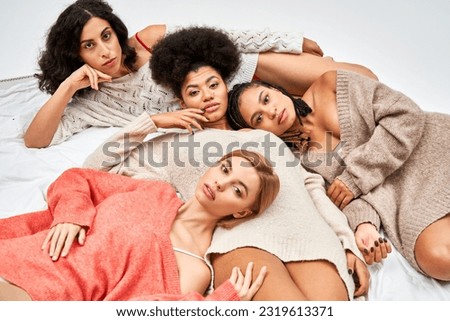 High angle view of multiethnic group of women in stylish warm jumpers looking at camera while lying on bed isolated on grey, different body types and self-acceptance, multicultural representation