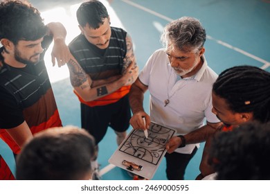 High angle view of a multiracial male basketball team and elderly coach in huddle while discussing game tactics. Planing the game tactic and teaching players defense. Diversity and teamwork. - Powered by Shutterstock