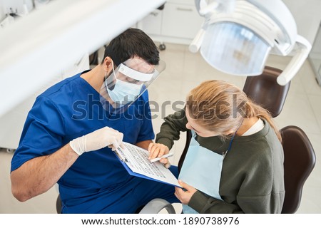 High angle view of multiracial man stomatologist having conversation with his female patient and filing documents while sitting at the dentistry room. People, medicine, stomatology and health care