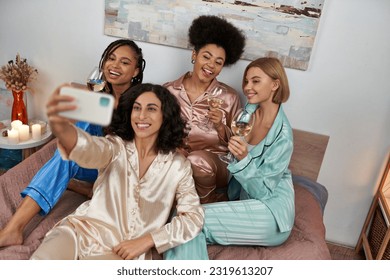 High angle view of multiracial woman in pajama taking selfie with cheerful multiethnic girlfriends with wine and sitting on bed at home, bonding time in comfortable sleepwear, slumber party - Powered by Shutterstock