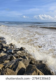 High angle view of ocean waves crashing against heavy granite boulders on a coastal sea wall under a blue sky