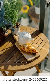 High angle view on ceramic teapot, fresh baked croissant on wooden round table on balcony. Summer terrace with simple garden furniture and breakfast on top. Perfect morning in coffee shop
