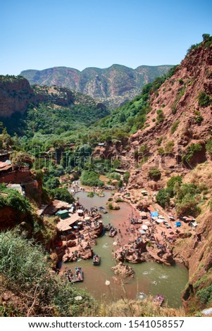 High angle view on red mountain gorge with water, green tropical park and vacationing peole below shot at Ouzoud Cascades, Morocco