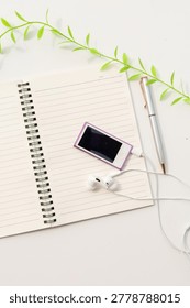 High angle view of open blank lined spiral notebook with pink digital music player, earbuds, and pen framed by green leaves