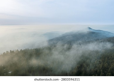 A high angle view over Hoher Bogen in the Bavarian Forest, Germany, shows rolling forested mountains partially covered by low-lying fog and mist. Dense evergreen woodland stretches across the scene while a distant summit with a radio tower emerges from the haze under a pale cloudy sky. The soft, natural lighting and layered hills create an atmospheric mountain landscape with ample copy space in the sky.