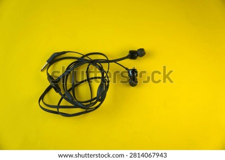 High angle view of a pair of black wired in-ear earphones with a tangled cable and a 3.5mm audio jack connector, isolated on a bright yellow background. Concept of chaotic cable mess, personal audio technology, and music listening.