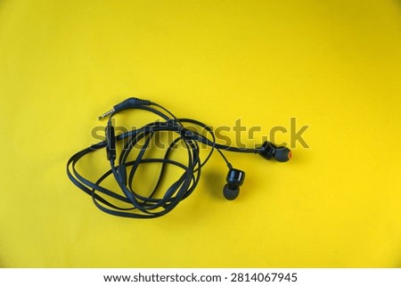 High angle view of a pair of black wired in-ear earphones with a tangled cable and a 3.5mm audio jack connector, isolated on a bright yellow background. Concept of chaotic cable mess, personal audio technology, and music listening.