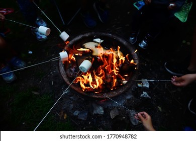 High angle view of people roasting marshmallows on skewers over fire pit at campsite. Enjoying summer outdoor camping fun and friend togetherness when park and campsite reopen after pandemic lockdown.