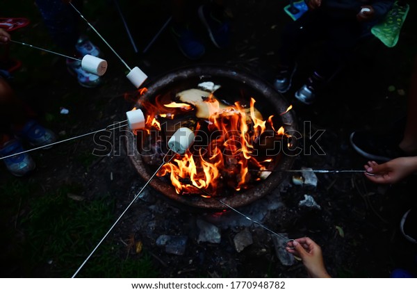 High angle view of people roasting marshmallows
on skewers over fire pit at campsite. Enjoying summer outdoor
camping fun and friend togetherness when park and campsite reopen
after pandemic lockdown.