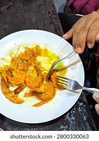 High angle view of a person using a fork and spoon to eat delicious cooked prawns in yellow sauce on a white plate.