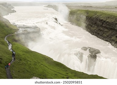 High angle view of powerful Gullfoss Waterfall in Iceland with small figures of people enjoying view from rocky cliff and footpath with visitors walking there