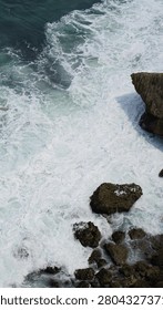 High angle view of powerful ocean waves breaking against coastal cliffs and creating thick white sea foam.