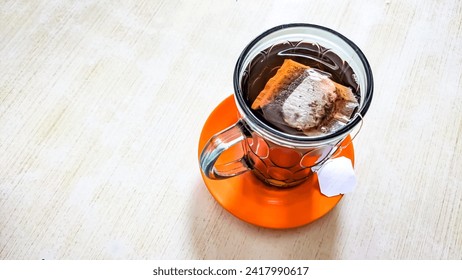 High angle view. A presentation hot drink in the form of a tea bag in a glass with a red plastic saucer on a wooden-patterned plywood table. One of a popular beverage service local indonesian culinary