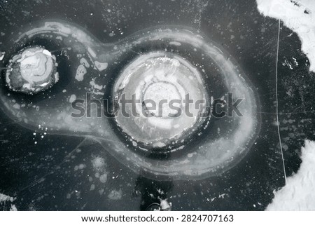 High angle view of rare circular ice formations and trapped methane bubbles frozen inside dark blue pond ice in winter. Concentric circular ice rings and frozen air bubbles forming an abstract figure eight pattern on dark lake ice framed by white snow