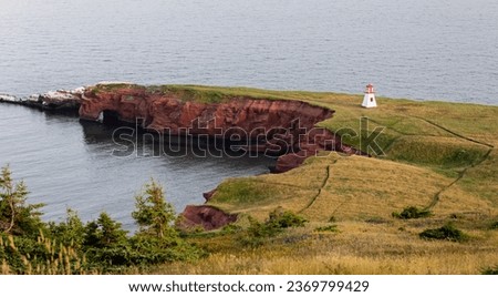 high angle view of a red rock cliff and a terrain with grass and a small white and red lighthouse on the side of the sea
