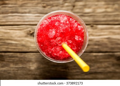 High Angle View Of Refreshing And Cool Frozen Red Fruit Slush Drink In Plastic Cup Served On Rustic Wooden Table With Bright Yellow Straw And Copy Space