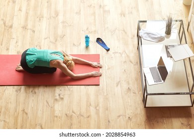 High Angle View Of Relaxed Mature Woman Practicing Yoga At Home, Sitting In Child Pose On A Mat In Front Of Laptop Monitor