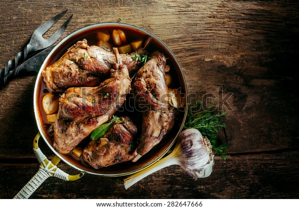 High Angle View of Roasted Rabbit Haunches in Pan\
with Stewed Vegetables on Rustic Wooden Table Surface with Bulb of\
Garlic and Copy Space