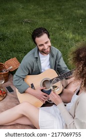High Angle View Of Romantic Man Playing Acoustic Guitar Near Curly Woman With Glass Of Wine During Picnic