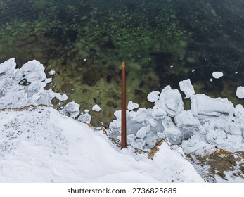 High angle view of a rusty iron pipe protruding from a snowy cliff over the clear Baltic Sea water, Estonia.