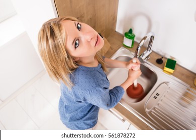 High Angle View Of Sad Young Woman Using Plunger In Blocked Kitchen Sink At Home