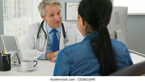 High Angle View Of Senior Caucasian Male Doctor In His Office Talking To New Patient And Smiling. Primary Care Physician Explaining Medical Issue To Woman