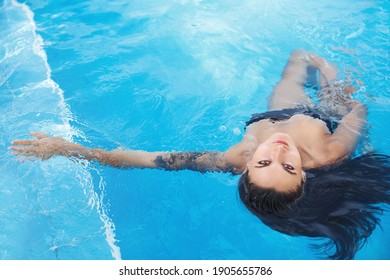 High angle view of sexy brunette woman lying on water in swimming pool of spa hotel, looking seductive at camera. Female model relaxing in poolside.