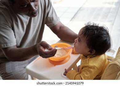 High angle view shot of mature African American man sitting in front of his baby daughter feeding her with puree - Shutterstock ID 2145941079