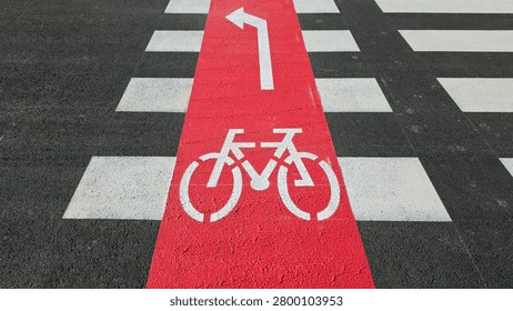 A high angle view showing a red painted bike lane crossing a black asphalt street with white pedestrian zebra stripes, featuring a stenciled bicycle icon and a sharp left turn directional arrow