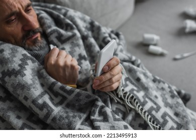 High Angle View Of Sick Man Wrapped In Warm Blanket Looking At Mobile Phone