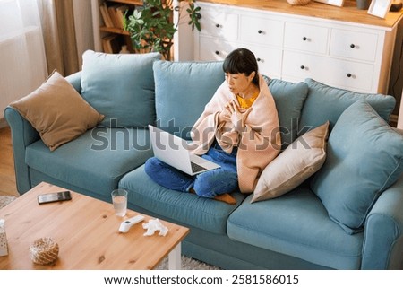 High angle view of sick middle aged Japanese woman sitting on living room couch covered with blanket having online doctor's appointment using laptop computer