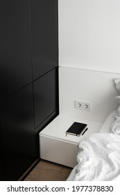 High angle view of side table with black notepad and pen near bed with white blanket. Part of bedroom interior with modern furniture, wardrobe and morning light in room, vertical shot
