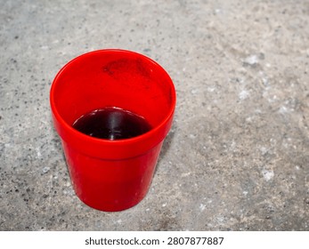 High angle view of a simple red plastic cup containing dark remaining coffee or liquid on textured grey concrete ground surface