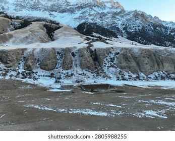 High angle view of a small, abandoned wooden building or cabin sitting on a barren plain beneath steep, rugged, snow-covered mountain slopes