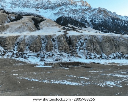 High angle view of a small, abandoned wooden building or cabin sitting on a barren plain beneath steep, rugged, snow-covered mountain slopes