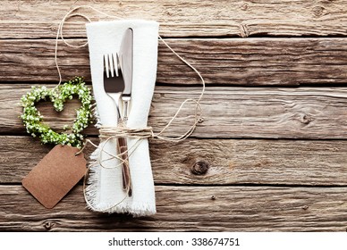 High Angle View Of Small Heart Shaped Greenery Wedding Wreath At Table Setting With Silver Knife And Fork Tied With String To Fringed White Napkin With Blank Tag On Rustic Wooden Table Surface