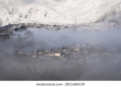  High angle view of a small mountain village partially obscured by dense morning fog and mist with snow-covered peaks in the background. Serene winter landscape of a rural settlement nestled in a valley, with thick clouds of fog rolling over the houses and majestic snowy mountains. Scenic view of Stepantsminda village (Kazbegi) in Georgia during winter, featuring traditional architecture under a blanket of mist and snow