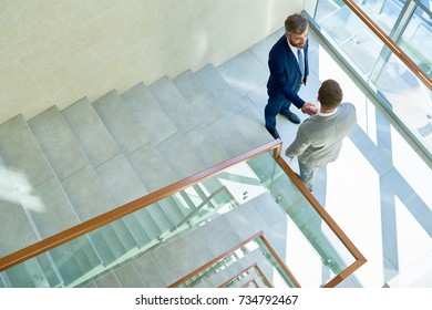 High Angle View Of Smiling Bearded Businessman Greeting His Young Colleague With Handshake While Standing On Staircase Landing Of Modern Office Building