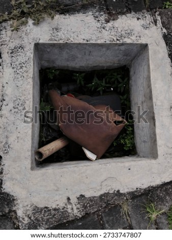 High angle view of a square concrete drainage hole filled with organic waste and plastic trash on a stone pavement outdoors during the day