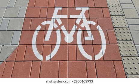A high angle view of a stenciled white bicycle icon on a reddish brown interlocking brick lane flanked by grey stone pavers and textured tactile paving for the visually impaired