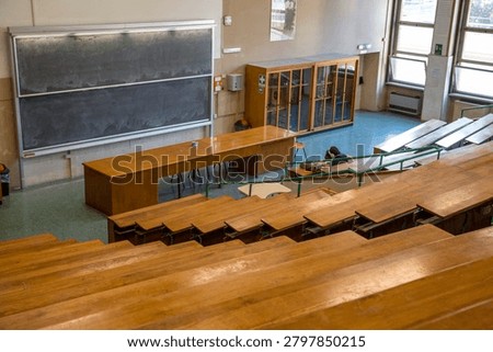High angle view of a student sitting alone in a large university lecture hall with rows of wooden desks, facing a massive double blackboard and a long wooden professor desk in an old classroom.